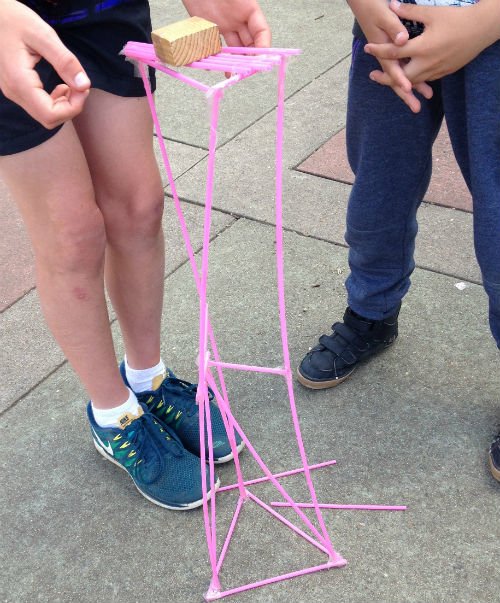 Straw tower building challenge, with pink straws taped together to raise a wooden block above a concrete surface 