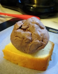 Ball of brown ice cream on top of a rectangular slice of yellow cake, sitting on a sheet of white baking paper with red spatula in background
