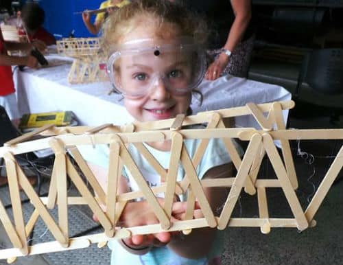 Building bridges using paddle pop wooden sticks. A girl in this photo is wearing safety glasses and smiling whilst holding up her bridge design