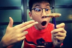 Boy with black rimmed glasses looks in awe , one hand pointing at other hand holding a pencil with a fidget spinner balanced on top