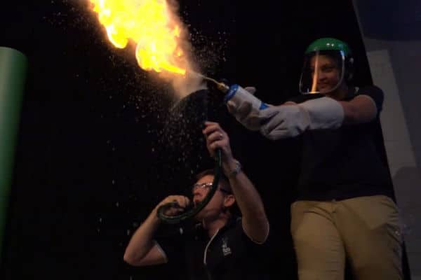 Holly from Fizzics wearing face shield and gloves while holding blow torch with Ben crouched next to her blowing flour through a hose and funnel making a large yellow fireball