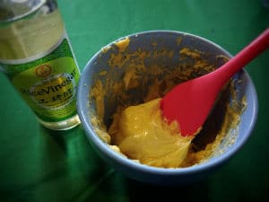 bottle of rice vinegar next to blue bowl of yellow mayonnaise with red spatula sticking out of it all on green tablecloth