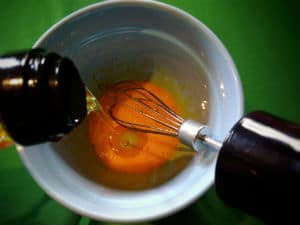 Whisk in the egg yolks in a blue bowl as oil pours in, all on green tablecloth