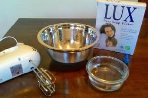 A box of lux soap flakes, a large metal bowl, a small glass bowl with water and a white electric beater on a brown table