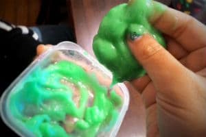 A hand full of green slime with a plastic container of green slime in the background