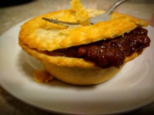 Meat pie yellow pastry brown mince and gravy oozing out of edge of pie as silver fork pushed down on top, on white plate