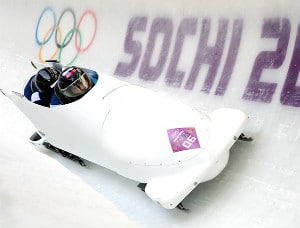 Two people wearing black helmets in a white bobsleigh on a track with Olympic games logo and sochi