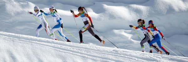 5 female olympic cross country skiers skiing uphill in white snow white clouds in the background