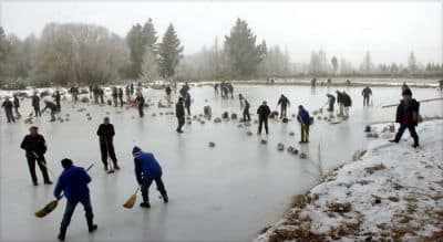 Numerous people holding brooms on an ice lake playing curling with granite stones, snow on the banks and misty trees in the background in naseby