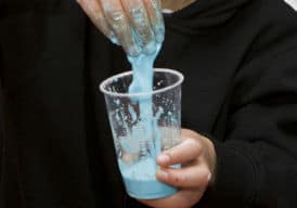 One hand holding a clear plastic cup with green slime inside, the other hand covered with dripping sline