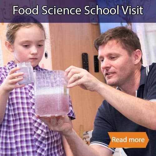 A student holding a plastic container whilst watching a purple solution fizz in a larger container (held by a teacher)